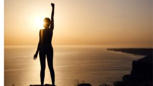 Figura de mujer alzando el brazo en señal de victoria mientras contempla un amanecer en la cima de una montaña, simbolizando el empoderamiento y la autoconfianza.