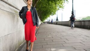 Mujer caminando con confianza en un paseo arbolado, vistiendo un vestido rojo y una chaqueta negra, proyectando seguridad y estilo.