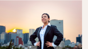 Mujer en traje de negocios con postura de poder, manos en la cintura y sonrisa segura, con un fondo de ciudad al atardecer que simboliza liderazgo y éxito profesional.